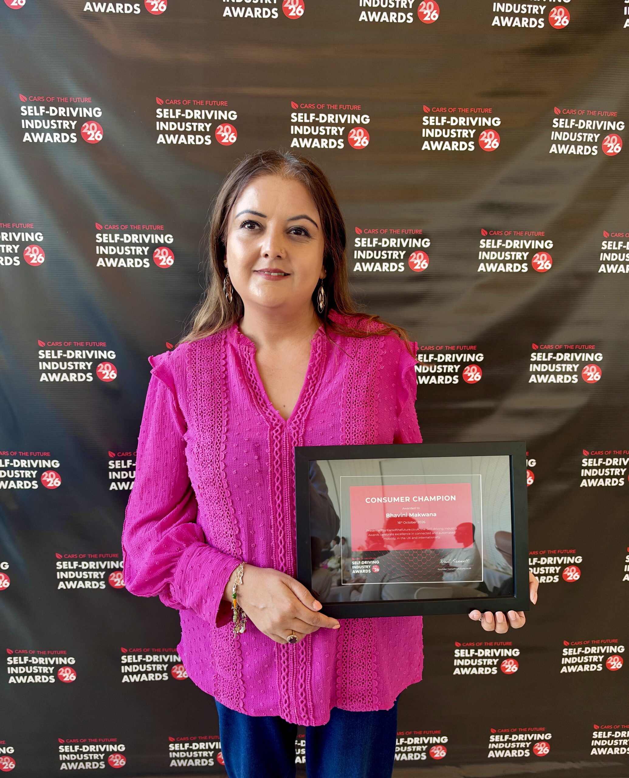 Bhavini smiling and holding her Consumer Champion framed award certificate in front of a media wall featuring the "Self-Driving Industry Awards 2026" logo pattern.