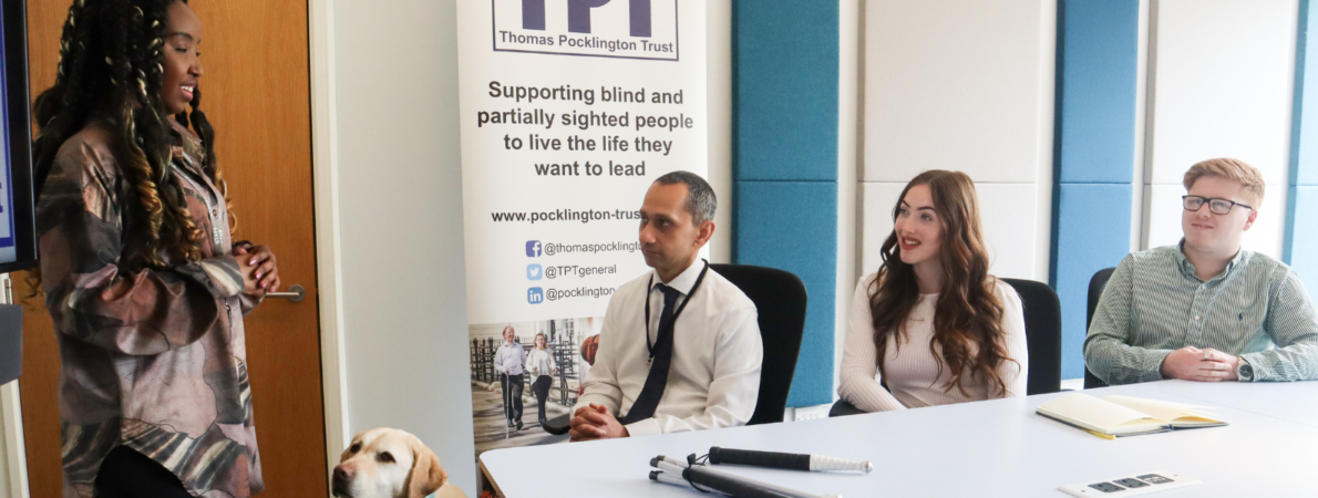 An image of three people sitting around a table and listening to a young woman standing and giving a presentation. There is a guide dog standing next to her and TPT's banner behind them. TPT and RNIB log on bottom left