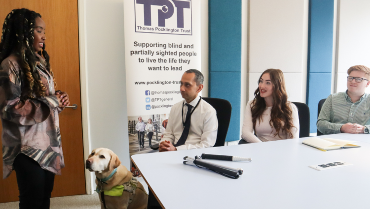 An image of three people sitting around a table and listening to a young woman standing and giving a presentation. There is a guide dog standing next to her and TPT's banner behind them. TPT and RNIB log on bottom left | links to article: Test Event