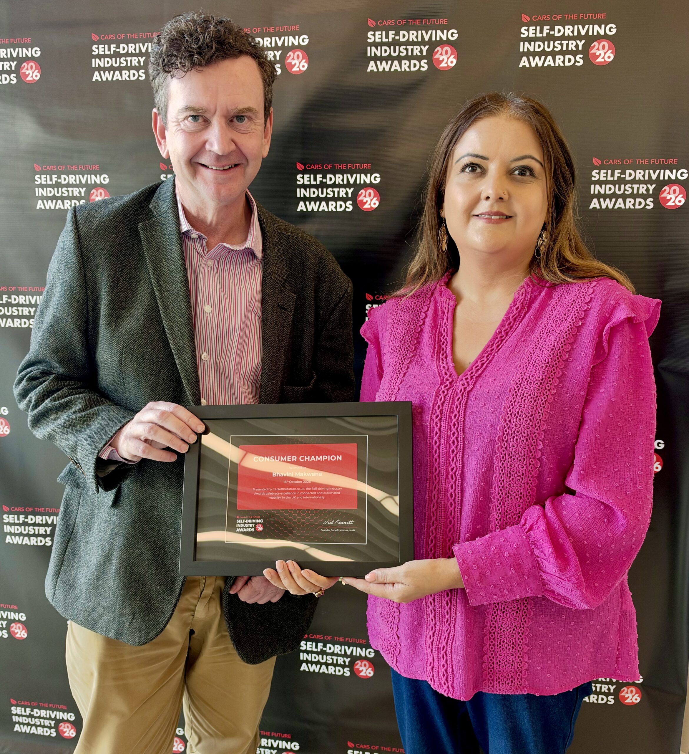 Bhavini, Consumer Champion award winner, standing alongside Charles, Chief Executive Officer at Thomas Pocklington Trust, in front of the media wall at the Self-Driving Industry Awards 2026.